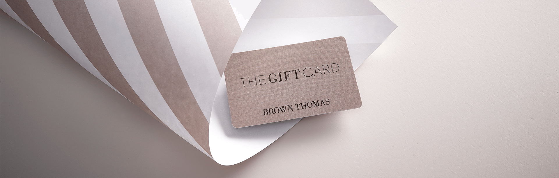 A Brown Thomas Gift Card rests on elegant wrapping paper, featuring muted striped patterns on a soft background.